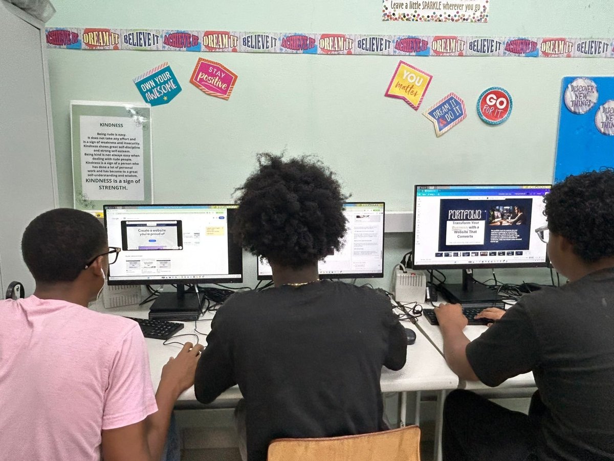 Three students working at computers in a classroom decorated with colorful posters on the wall