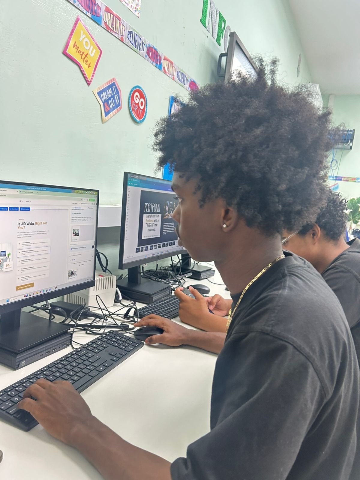 Student with curly hair working at a desk with dual monitors in a colorful classroom with decorations on walls